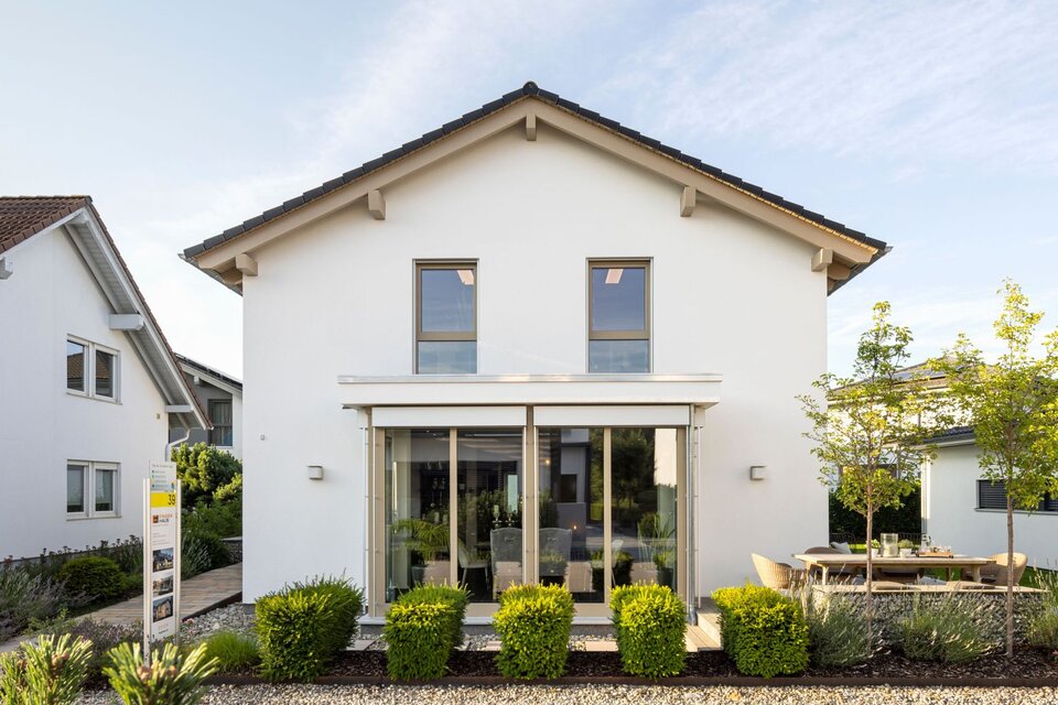 Frontansicht des Musterhauses MEDLEY 300 B in Poing mit Wintergarten-Erker und moderner Gartenanlage.