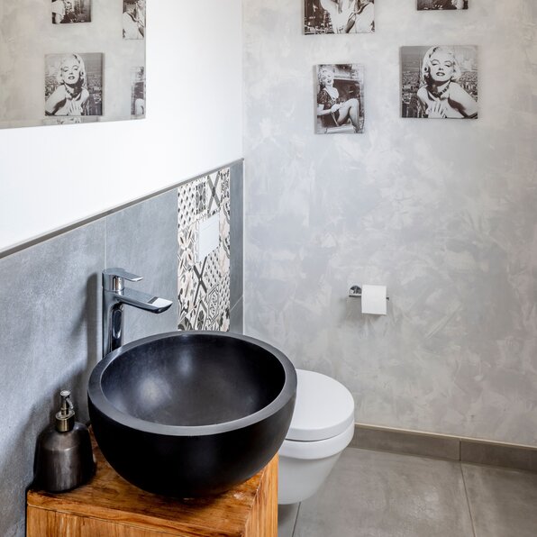 Gäste-WC mit schwarzem rundem Aufsatzwaschbecken auf einem Holzunterschrank, weißer Toilette, großem Spiegel und grauen Wand- und Bodenflächen.
