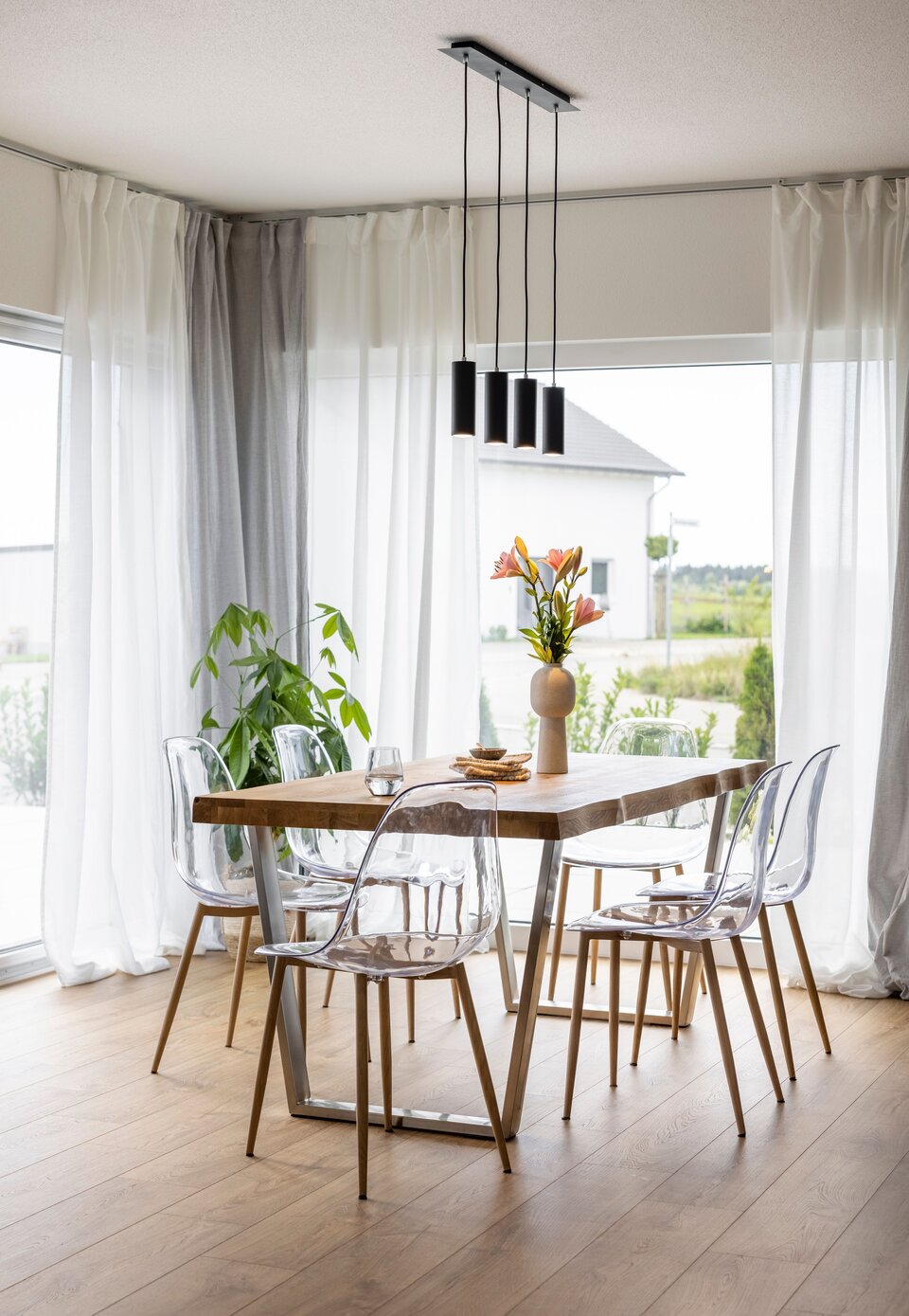 Helles Esszimmer mit großen Fenstern, Vorhängen und rundem Holztisch; freundliche und luftige Raumatmosphäre.