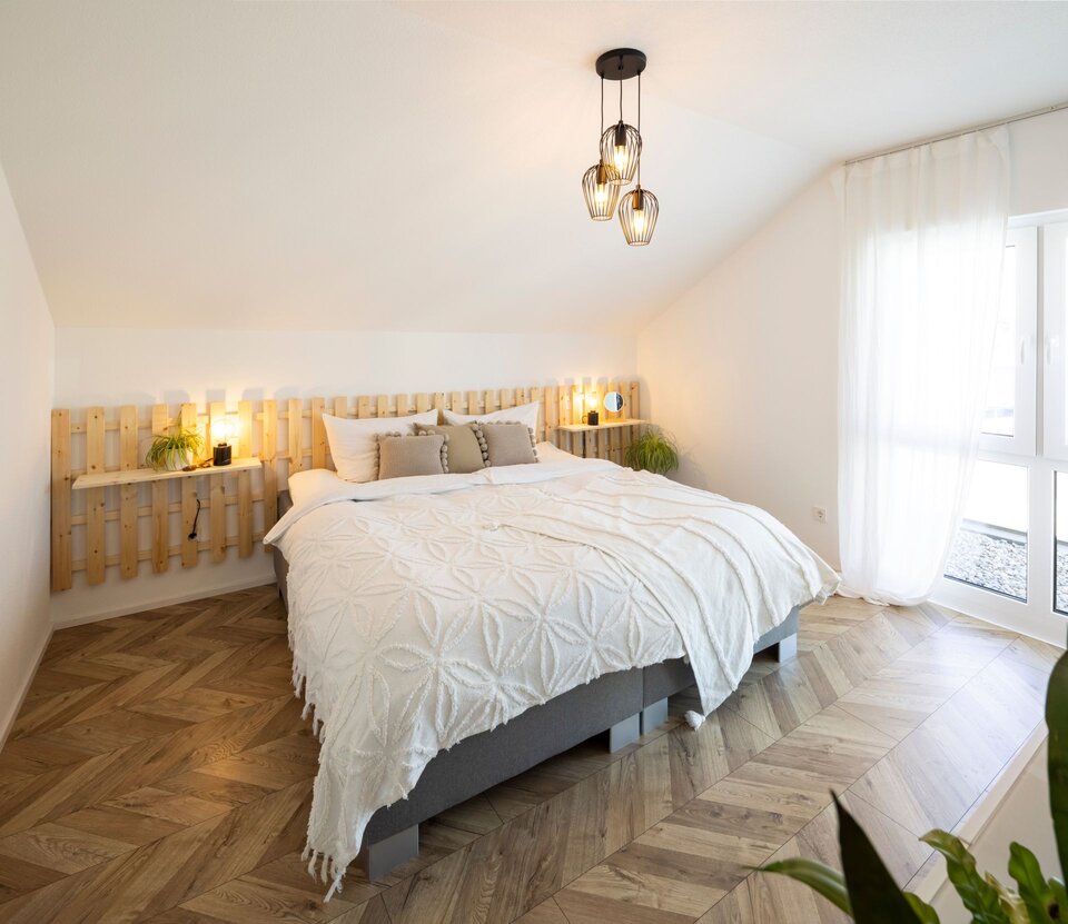 Schlafzimmer mit Doppelbett, Holzrückwand und Dachschräge. Helle Bettwäsche, Holzfußboden und Fenster sorgen für gemütliches Licht.