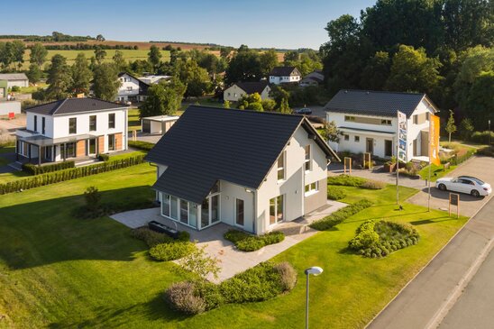 Luftaufnahme des Musterhausparks von FingerHaus in Frankenberg mit mehreren modernen Musterhäusern, gepflegten Grünflächen und Flaggen. Im Hintergrund sind Felder, Bäume und Wohnhäuser der Umgebung zu sehen.