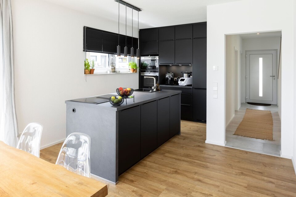 Moderne Küche mit dunklen Fronten, heller Arbeitsplatte und transparenten Stühlen am Esstisch. Klare Linien, Holzboden und Tageslicht durch große Fenster.