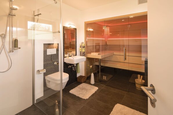 Modernes Badezimmer mit Dusche, WC, Waschbecken und angrenzender Sauna hinter Glas, in warmem Licht eines FingerHaus-Hauses.