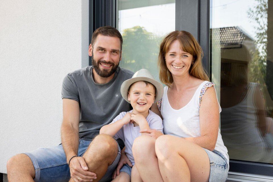 Familie mit zwei Erwachsenen und einem Kind sitzt vor einer Terrassentür und blickt lächelnd in die Kamera