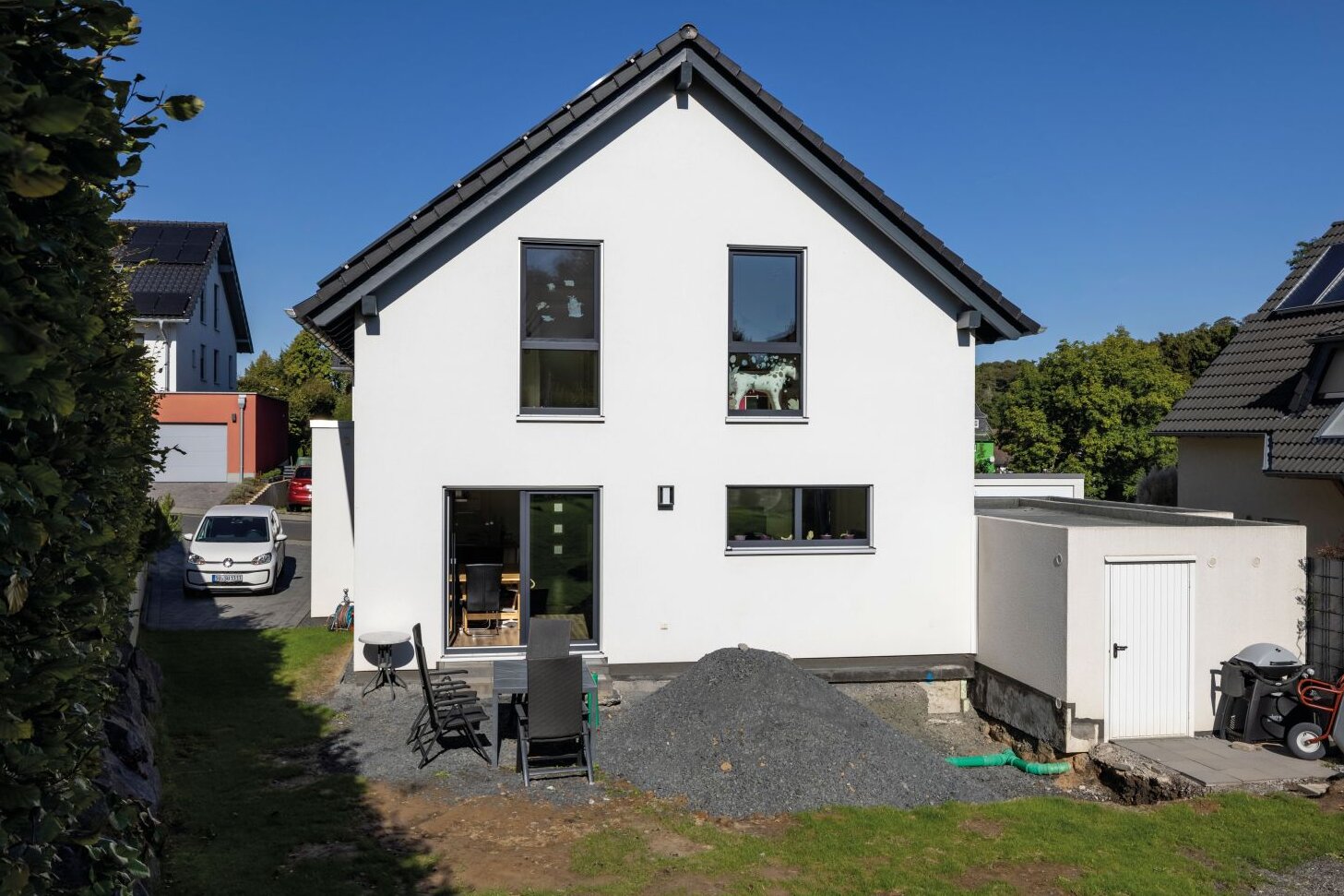 Rückansicht eines Einfamilienhauses mit weißer Fassade und dunklem Satteldach während der Gartengestaltung. Im Garten liegen Erd- und Schotterhaufen, Gartenmöbel und Werkzeuge stehen bereit.