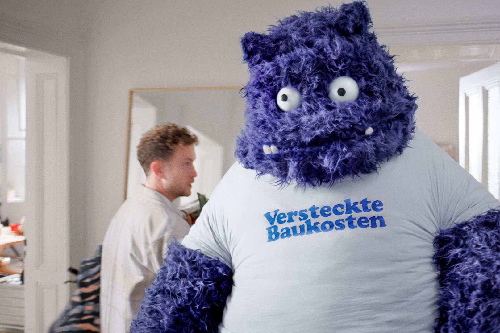 Ein großes blaues, plüschartiges Wesen mit der Aufschrift „Versteckte Baukosten“ auf dem T-Shirt steht in einer Wohnung – Symbolfigur aus der FingerHaus-Kampagne „Das Unerwartete“, die typische Sorgen beim Hausbau thematisiert.