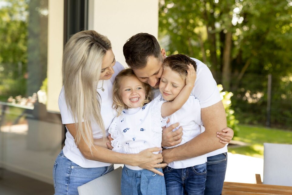 Eine glückliche vierköpfige Familie – Mutter, Vater, Tochter und Sohn – umarmt sich lachend auf der Terrasse eines modernen FingerHaus-Hauses an einem sonnigen Tag.