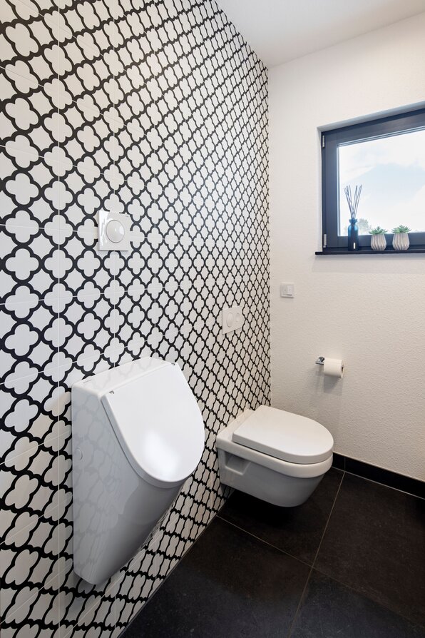 Modernes Gäste-WC mit auffälliger schwarz-weißer Musterfliese, Wand-WC, Urinal und kleinem Fenster mit Dekoration.