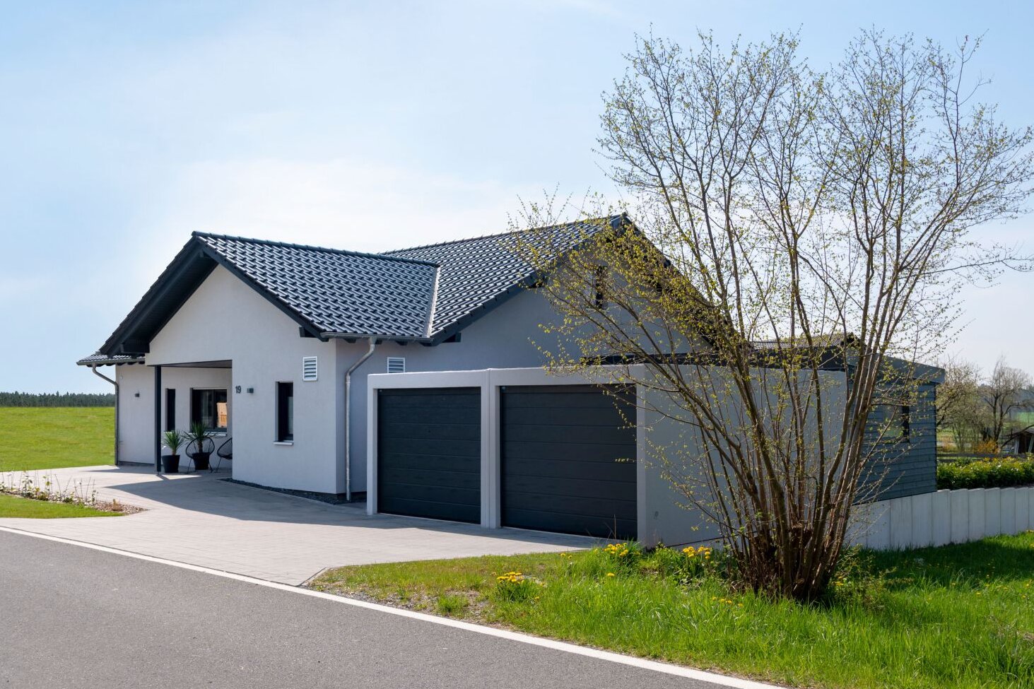 Außenansicht eines modernen FingerHaus NIVO 130 mit heller Fassade, dunklem Satteldach, überdachtem Eingangsbereich und großer Doppelgarage, gelegen an einer ruhigen Straße neben einer weitläufigen Wiese.