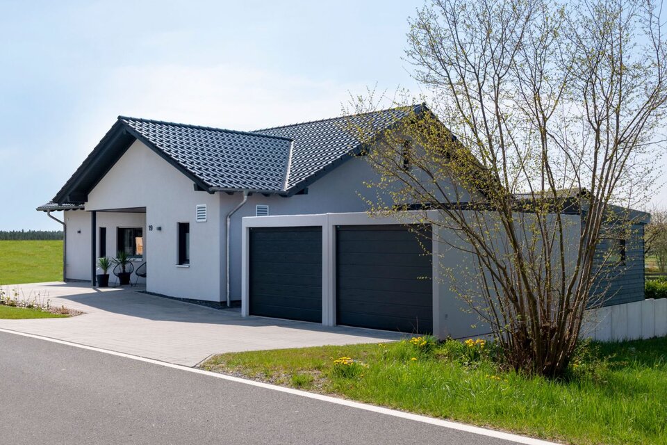 Außenansicht eines modernen FingerHaus NIVO 130 mit heller Fassade, dunklem Satteldach, überdachtem Eingangsbereich und großer Doppelgarage, gelegen an einer ruhigen Straße neben einer weitläufigen Wiese.