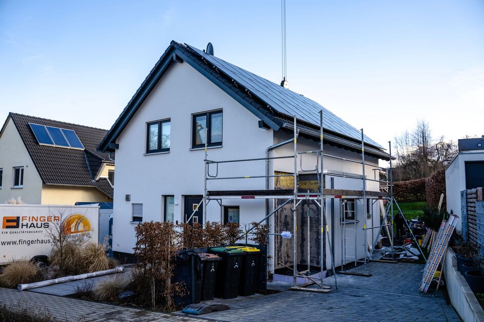 Einfamilienhaus während eines Umbaus mit Gerüst an der Fassade. Das Haus hat ein dunkles Satteldach und weiße Putzwände. Im Vordergrund stehen Mülltonnen, Baumaterial und ein FingerHaus-Schild.