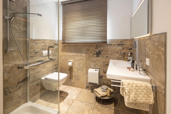 Badezimmer im Musterhaus VIO Köln mit bodengleicher Dusche, modernen Fliesen in Natursteinoptik und stilvoller Waschtischlösung.