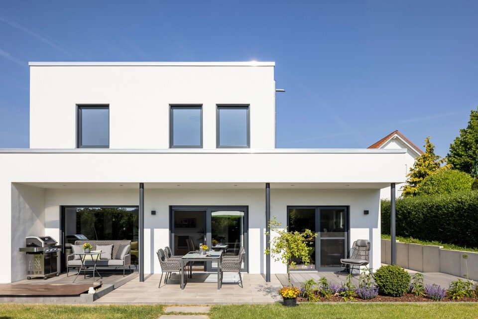 Außenansicht eines frei geplanten FingerHaus im modernen Bauhaus-Stil mit weißer Fassade. Überdachte Terrasse mit Sitzgruppe, Loungemöbeln und Grillplatz sowie gepflegtem Garten.