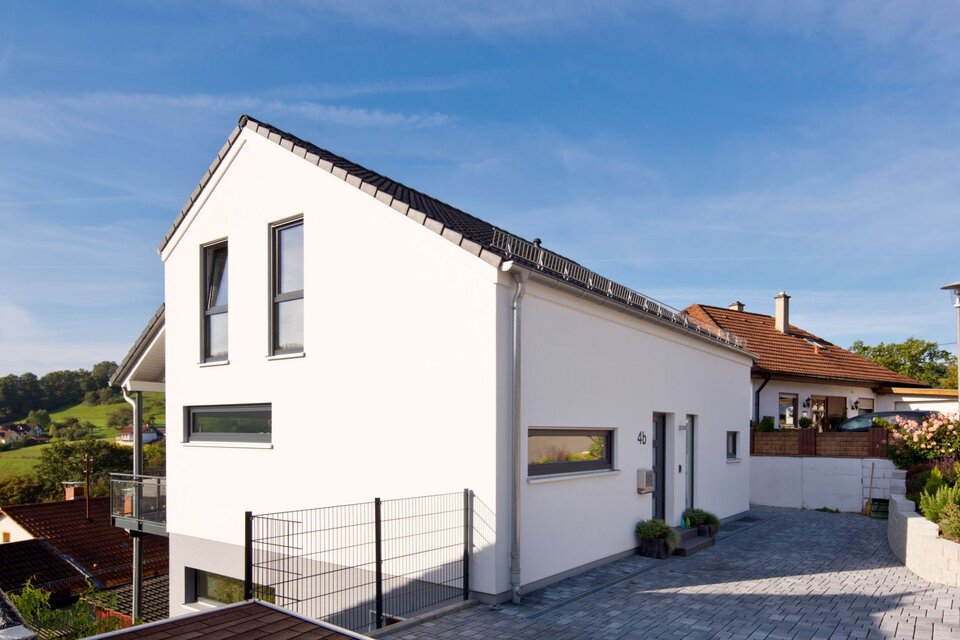 Modernes FingerHaus-Einfamilienhaus mit weißer Putzfassade, dunklem Satteldach, klarer Linienführung und Einfahrt, gelegen in Hanglage mit Aussicht ins Grüne.