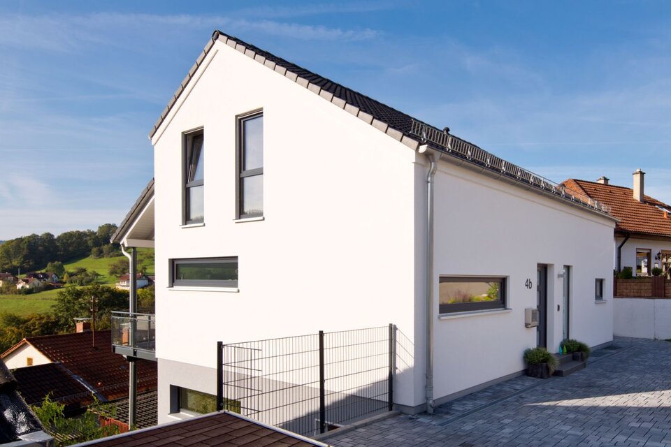 Modernes FingerHaus-Einfamilienhaus mit weißer Putzfassade, dunklem Satteldach, klarer Linienführung und Einfahrt, gelegen in Hanglage mit Aussicht ins Grüne.