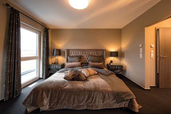 Gemütliches Schlafzimmer im FingerHaus MEDLEY 300 B Musterhaus Berlin/Werder mit Polsterbett, warmen Farben und dekorativen Kissen.