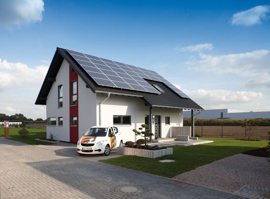 Fertighaus Musterhaus FingerHaus VIO 400 in Köln mit heller Fassade, rotem Fassadenelement und großer Photovoltaikanlage auf dem Dach, davor ein FingerHaus-Auto auf der gepflasterten Einfahrt.