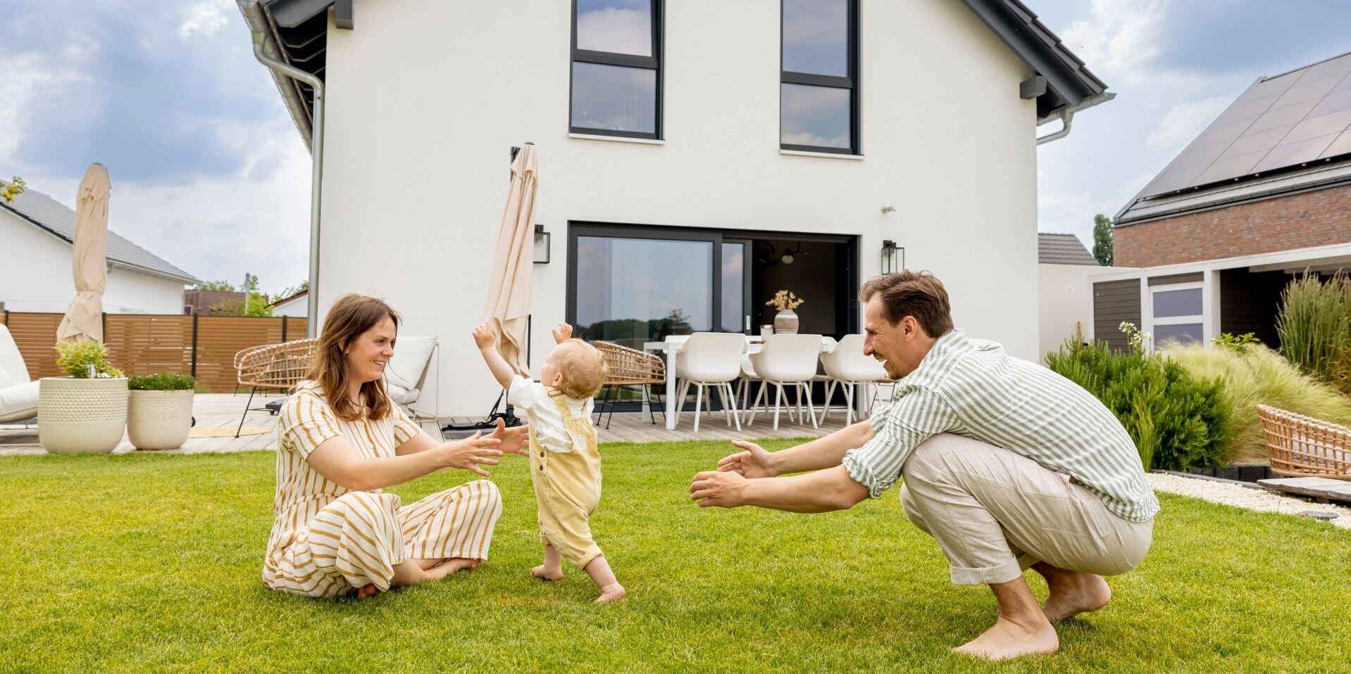 Eine junge Familie spielt barfuß auf dem Rasen vor ihrem modernen FingerHaus-Haus. Das Baby läuft lachend zwischen Mutter und Vater hin und her.