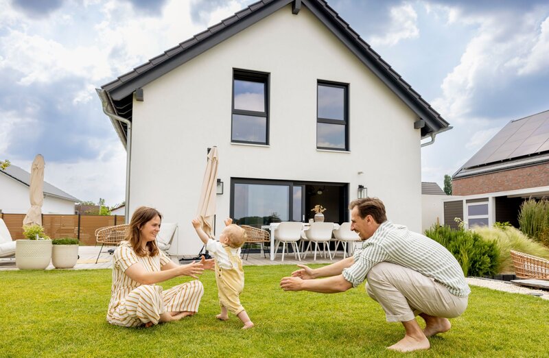 Eine junge Familie spielt barfuß auf dem Rasen vor ihrem modernen FingerHaus-Haus. Das Baby läuft lachend zwischen Mutter und Vater hin und her.
