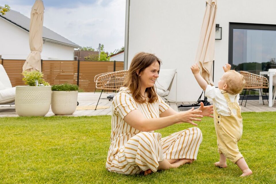 Eine junge Familie spielt barfuß auf dem Rasen vor ihrem modernen FingerHaus-Haus. Das Baby läuft lachend zwischen Mutter und Vater hin und her.