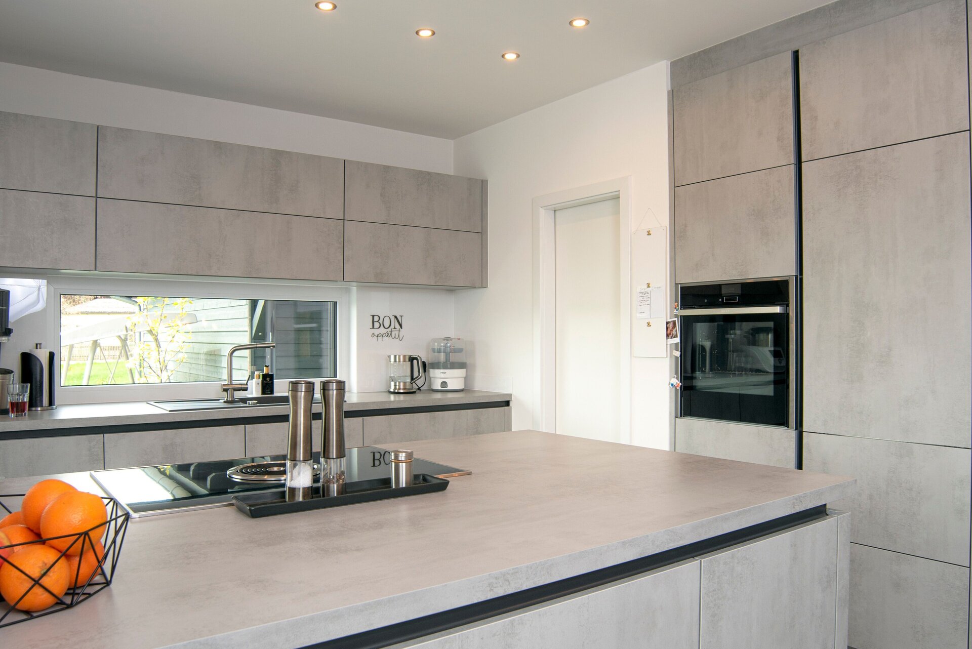 Moderne Küche in einem FingerHaus NIVO 130 mit grauen Fronten, großer Kochinsel, eingebautem Backofen und breitem Fenster über der Arbeitsfläche.