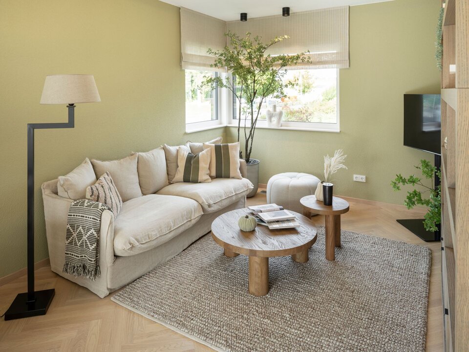 Gemütliches Wohnzimmer im Musterhaus JUNO 300 in Mülheim-Kärlich mit naturfarbenem Sofa, runden Holztischen und grünen Wänden.
