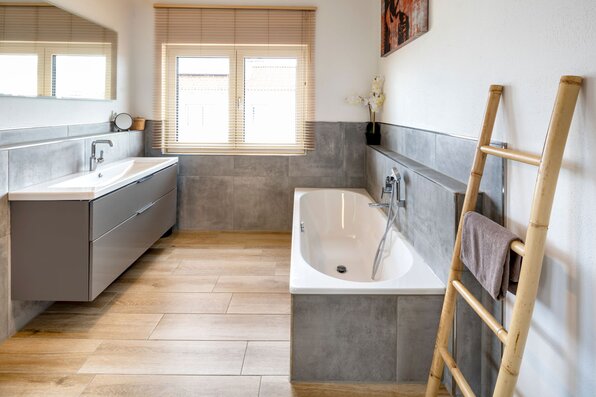 Badezimmer mit weißer Badewanne, grauem Waschtischunterschrank, großem Spiegel, grauen Wandfliesen und Bodenfliesen in Holzoptik.