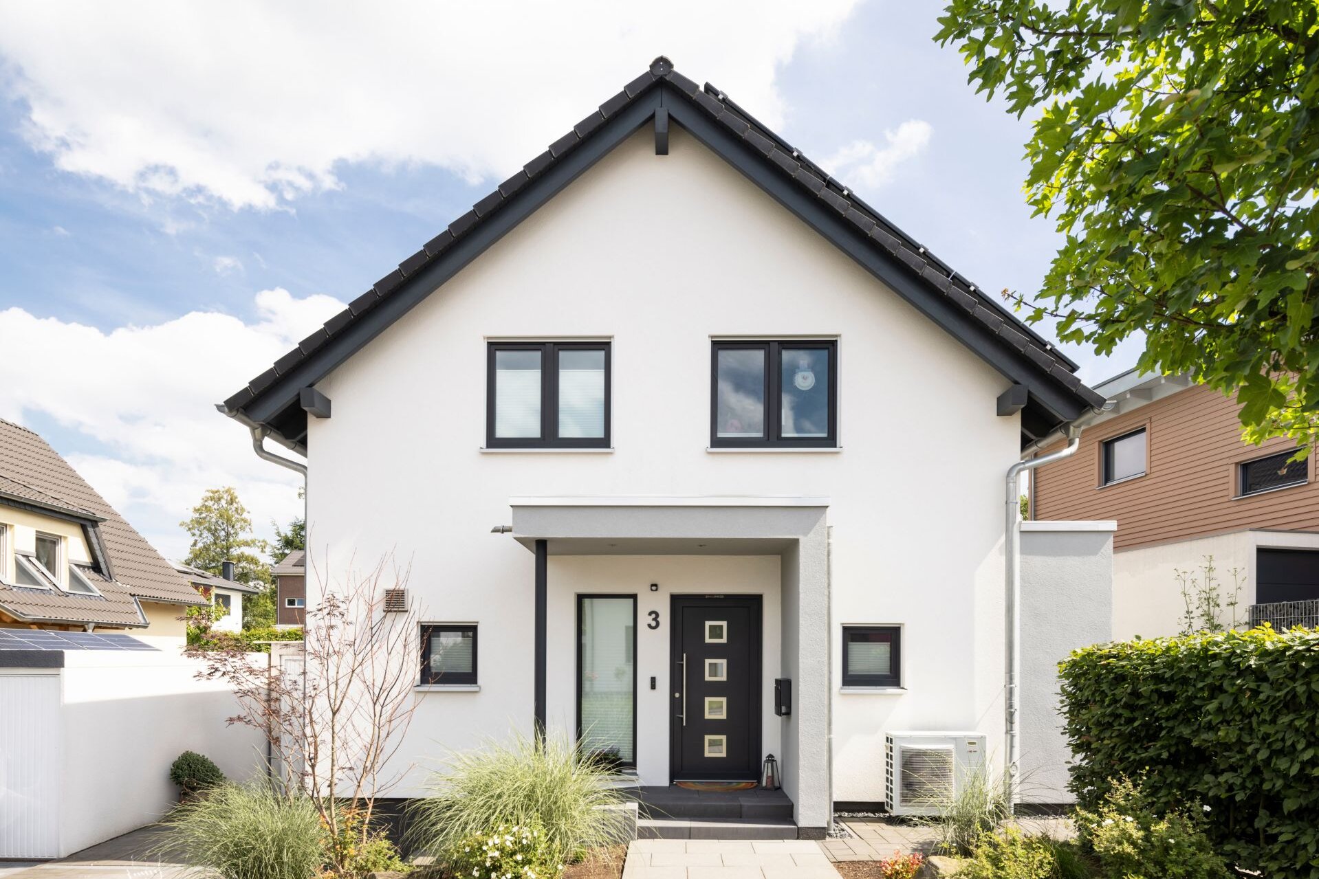 Frontansicht eines modernen Einfamilienhauses mit weißer Fassade, dunklem Satteldach und überdachtem Eingang. Ein gepflasterter Weg führt durch den gepflegten Vorgarten mit Blumen und Ziergräsern zur Haustür.