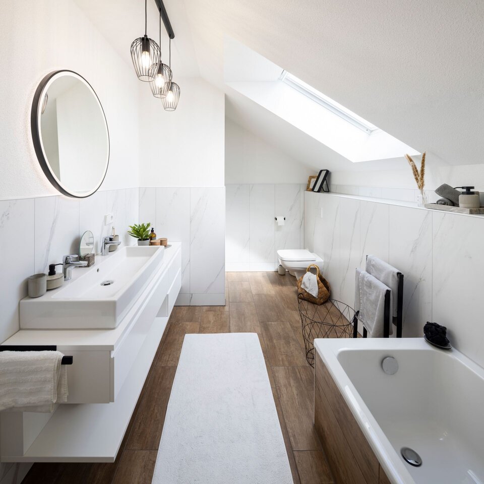 Modernes Badezimmer mit Waschtisch, großer Spiegelwand und Badewanne unter der Dachschräge. Helle Fliesen, Holzboden und dezente Beleuchtung.