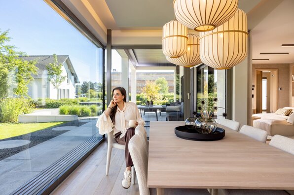 Food-Bloggerin Sally Özcan sitzt im Essbereich des Musterhauses NIVO »Sally Edition« im Musterhauspark Frankenberg mit einer Tasse in der Hand und Blick in den Garten.