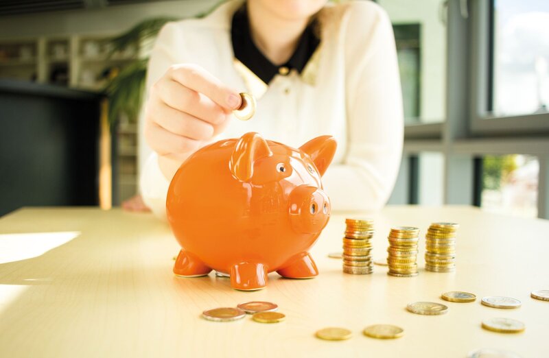 Hand wirft eine Münze in ein orangefarbenes Sparschwein, daneben liegen gestapelte Münzen auf einem Tisch