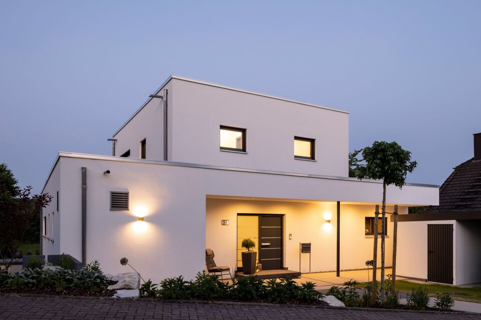 Überdachte Terrasse eines frei geplanten FingerHaus im modernen Bauhaus-Stil mit Sitzgruppe aus geflochtenen Stühlen und Tisch. Bodentiefe Fenster führen in den Wohnbereich, daneben gepflegte Bepflanzung.