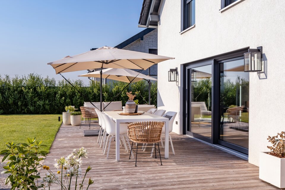 Große Holzterrasse eines modernen FingerHaus-Hauses mit stilvollen Gartenmöbeln, Sonnenschirmen und blühenden Pflanzen an einem klaren Sommertag.