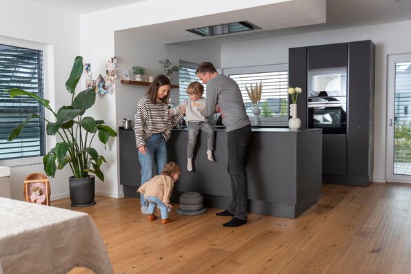 Eine Familie mit zwei kleinen Kindern steht in der modernen Küche eines FingerHaus-Hauses. Die Eltern helfen ihrem Sohn beim Kochen, während die Tochter neugierig zusieht.