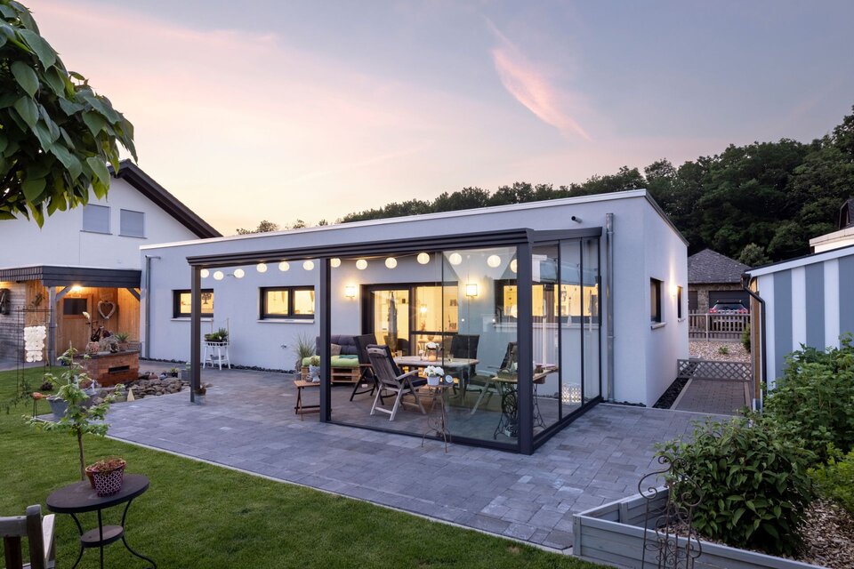 Modernes Flachdachhaus mit überdachter Glas-Terrasse, beleuchtetem Innenraum und gepflastertem Gartenbereich in der Abenddämmerung