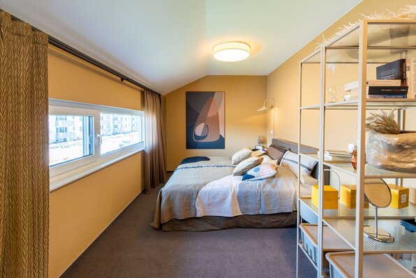 Gemütliches Schlafzimmer im FingerHaus Musterhaus TALO in Babenhausen mit Doppelbett, warmen Wandfarben und dekorativen Elementen. Moderne Einrichtung mit Regal, Kunst an der Wand und großen Fenstern für viel Tageslicht.