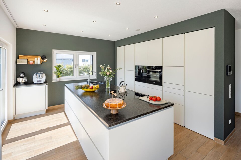 Moderne FingerHaus-Küche im MEDLEY 300 B mit großer Kochinsel, grün akzentuierter Wand und viel Tageslicht durch bodentiefe Fenster.