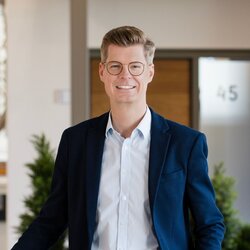 Lukas Tauschwitz, Marketingleiter bei FingerHaus, in blauem Sakko, weißem Hemd und beiger Hose lehnt mit einer Hand an einem Geländer in einem modernen Bürogebäude mit Pflanzen und Glastür im Hintergrund