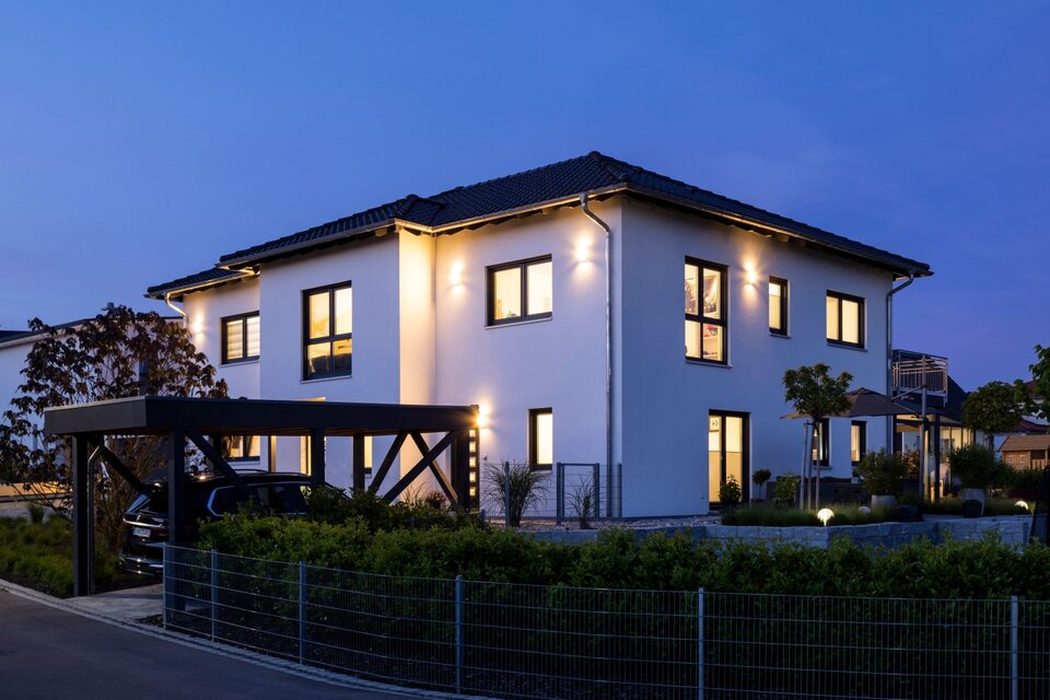 Modernes FingerHaus-Zweifamilienhaus in der Abenddämmerung mit eingeschalteter Außenbeleuchtung und Carport.