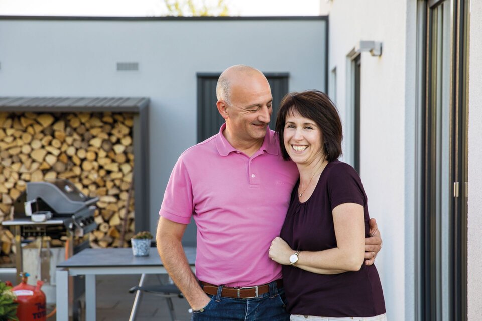 Glückliches Paar vor ihrem frei geplanten FingerHaus auf der Terrasse, im Hintergrund Holzlager und Grill.
