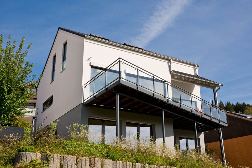 Modernes FingerHaus-Einfamilienhaus in Hanglage mit heller Fassade, großem Balkon mit Glasgeländer und bodentiefen Fenstern im Untergeschoss.