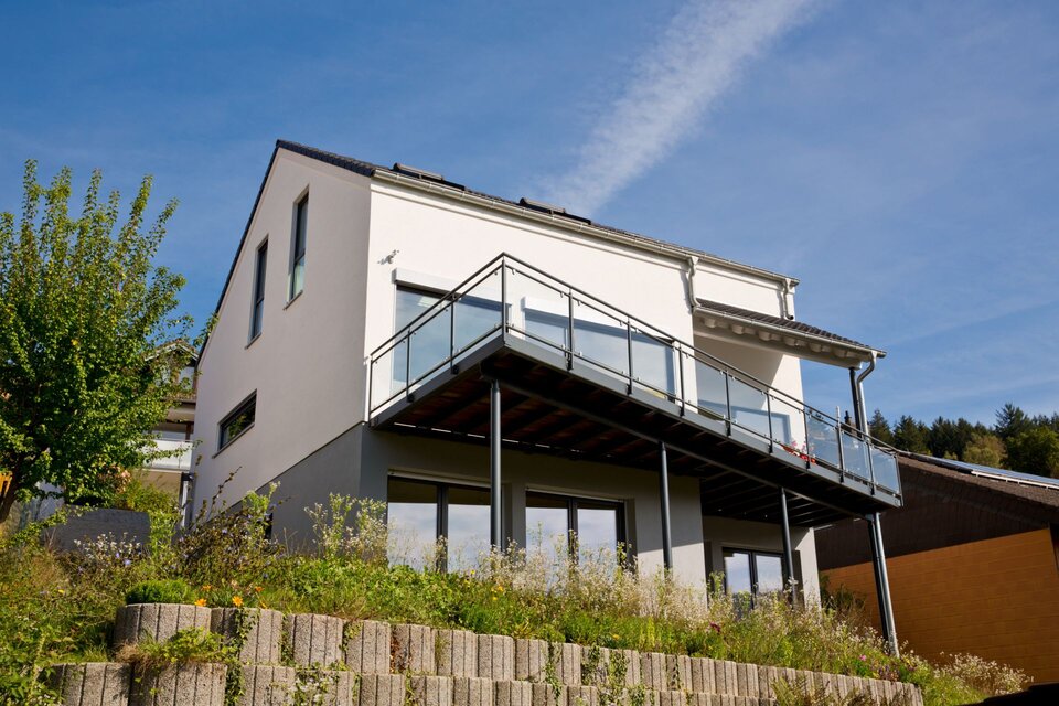 Modernes FingerHaus-Einfamilienhaus in Hanglage mit heller Fassade, großem Balkon mit Glasgeländer und bodentiefen Fenstern im Untergeschoss.