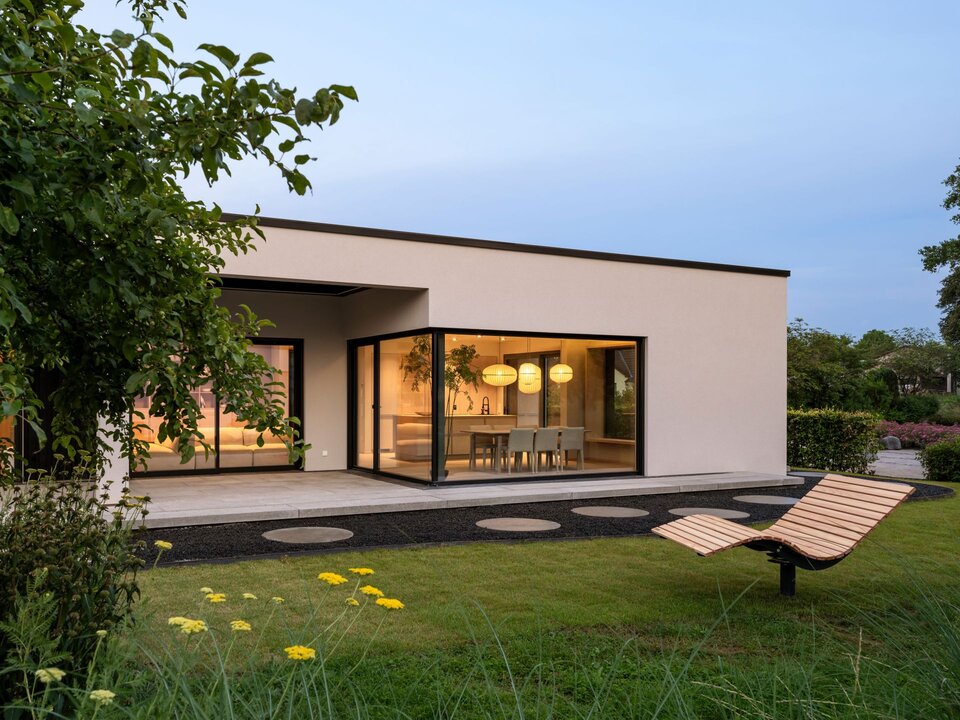 Terrassenansicht des Musterhauses NIVO in Frankenberg mit moderner Bungalowarchitektur, überdachter Terrasse, großzügigen Eckfenstern und beleuchtetem Wohn- und Essbereich in der Abenddämmerung.