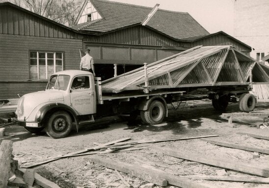 Schwarz-weiß-Fotografie eines Lastwagens mit einem großen, dreieckigen Holzfachwerk auf dem Anhänger, daneben ein Mann auf dem Fahrzeug, im Hintergrund ein Fachwerkhaus und eine Scheune.