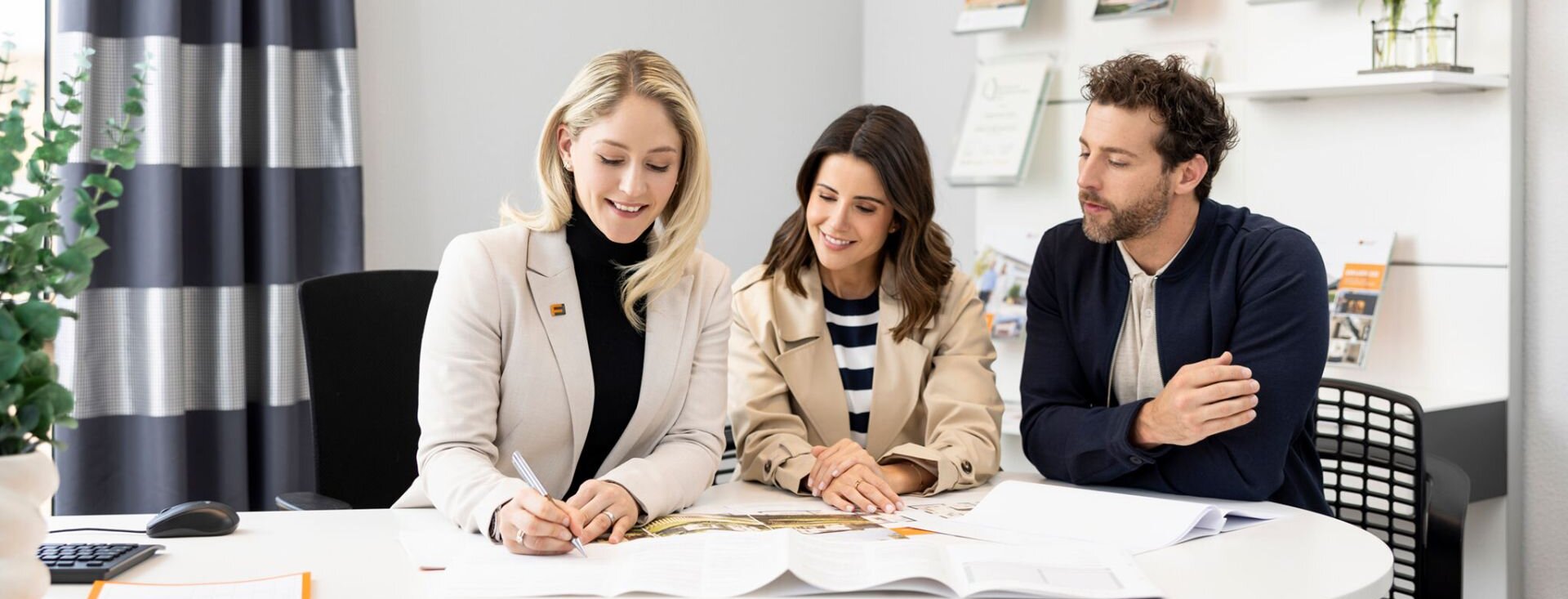 Interessentenpaar im ersten Beratungsgespräch mit einer FingerHaus-Fachberaterin, gemeinsam am Tisch bei der Durchsicht von Unterlagen und Hausplänen.