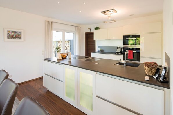 Moderne Küche in einem frei geplanten FingerHaus mit heller Kochinsel, eingebauten Elektrogeräten, dunkler Arbeitsplatte und Zugang zur Terrasse.