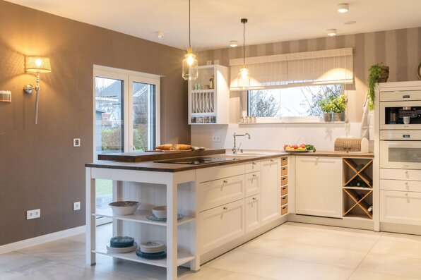 Helle Landhausküche im Musterhaus SENTO 503 in Bad Vilbel mit Kücheninsel, offenem Weinregal, modernen Einbaugeräten und gemütlicher Beleuchtung.