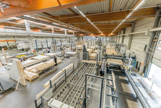 Blick in die moderne Fertigungshalle von FingerHaus mit automatisierten Produktionsanlagen, Holzrahmen-Elementen und Mitarbeitenden beim Zusammenbau von Fertighauswänden.