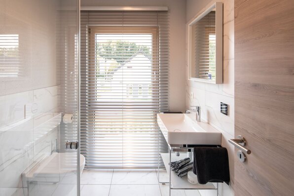 Badezimmer im FingerHaus-Musterhaus MEDLEY 300 B in Frankenberg mit bodengleicher Dusche mit Glaswand, wandhängendem WC, Waschtisch mit Aufsatzbecken, Spiegel und Fenster mit horizontalen Jalousien; helle Wand- und Bodenfliesen in Marmoroptik und Holzdekor.