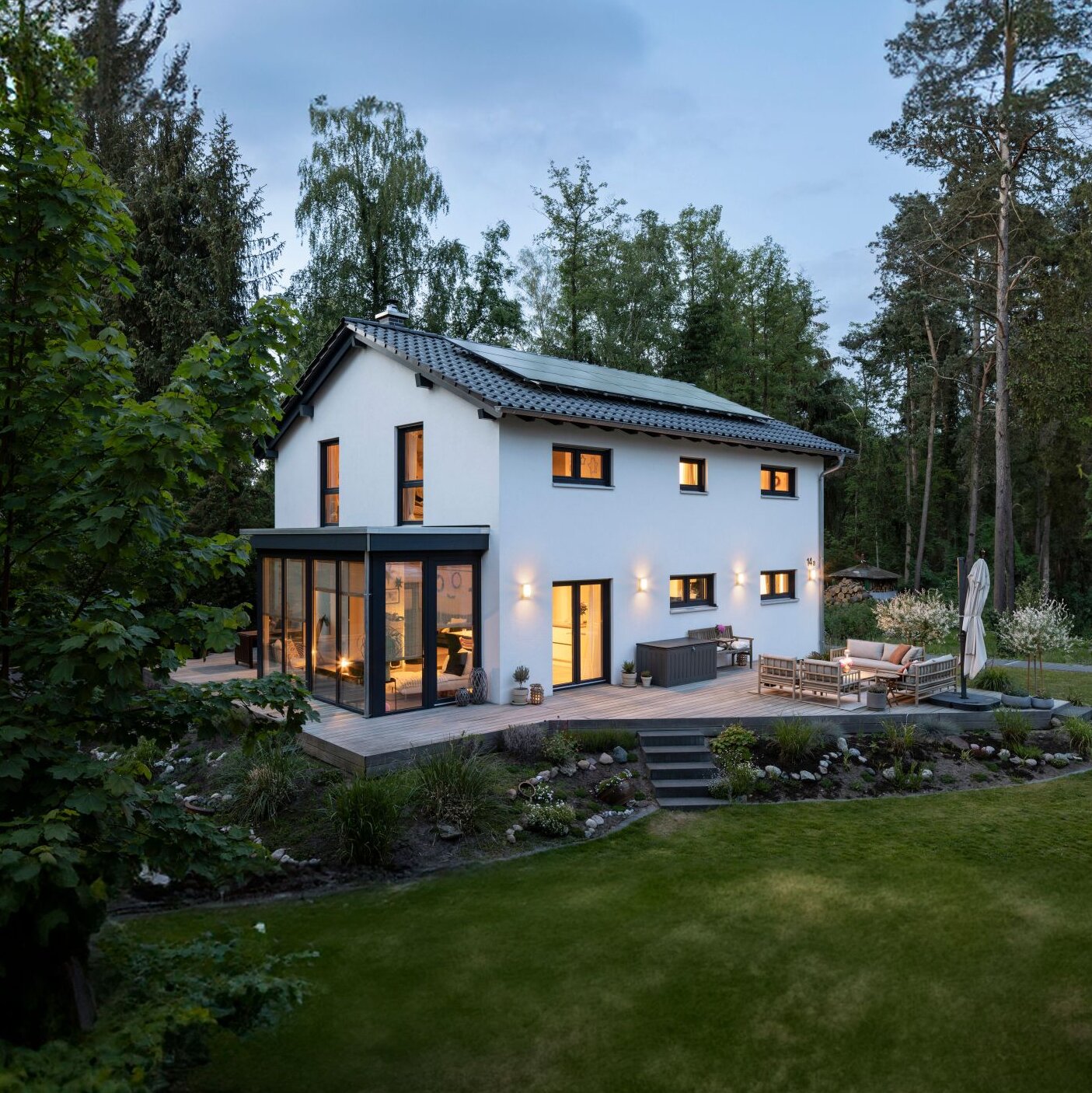 Zweigeschossiges Haus mit Satteldach und weißer Fassade, großem verglasten Anbau, Terrasse mit Sitzmöbeln sowie weitläufigem Garten, umgeben von hohen Bäumen in der Abenddämmerung.